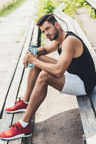young sportsman with bottle of water sitting on bench at sport playground 