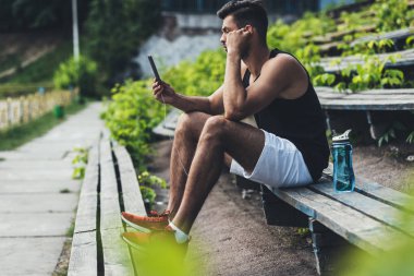genç sporcu spor Bahçesi, bankta yan görünüm şişe su akıllı telefon ve kulaklık ile müzik dinleme