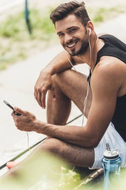 adam akıllı telefon ve kulaklık bankta ile müzik dinleme su şişe ile gülümseyen genç 