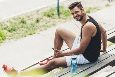 adam akıllı telefon ve kulaklık bankta ile müzik dinleme su şişe ile gülümseyen genç 