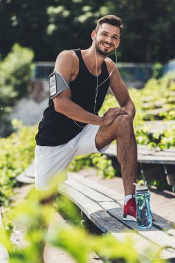 Kulaklıklar içinde mutlu sporcu ile çalışan spor Bahçesi, bankta egzersiz yaparak çantası içinde smartphone