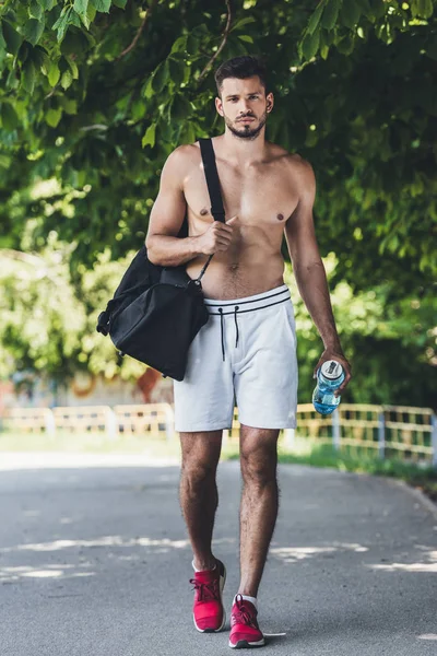 attractive young man with bag for sport equipment and fitness bottle walking under green tree