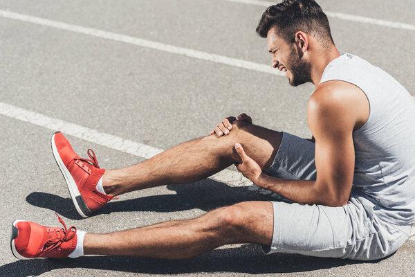athletic young runner with leg injury sitting on floor of running track