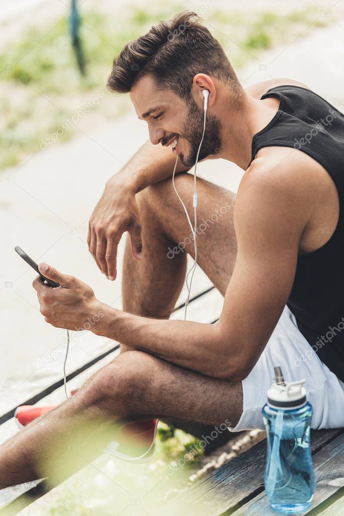 sportsman with bottle of water listening music with smartphone and earphones on bench at sport playground