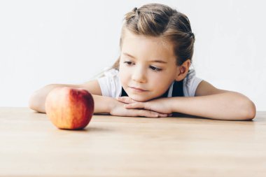 little schoolgirl sitting at table with red apple isolated on white clipart