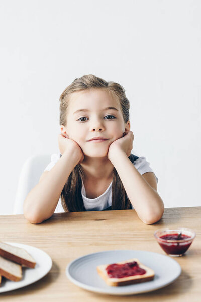little schoolgirl sitting at table and having toasts with jam for breakfast isolated on white