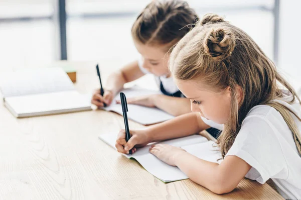 Piccole Studentesse Che Scrivono Quaderni Mentre Fanno Compiti Isolati Bianco — Foto Stock