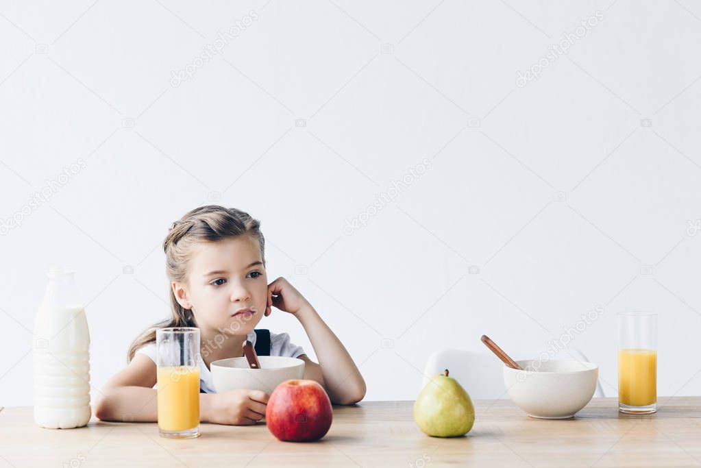 lonely little schoolgirl sitting at table during breakfast and looking away isolated on white