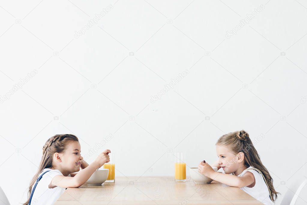 adorable smiling schoolgirls eating cereals with orange juice for breakfast while sitting in front of each other isolated on white