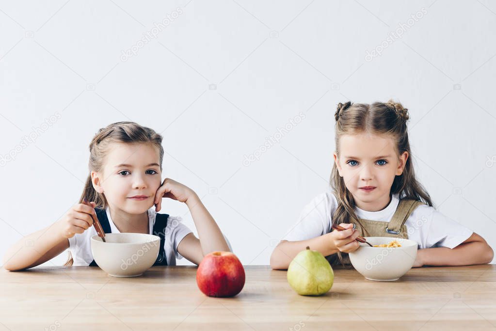 adorable smiling schoolgirls eating cereals with fruits for breakfast isolated on white