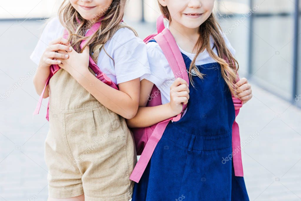 cropped shot of smiling schoolgirls with pink backpacks standing on street