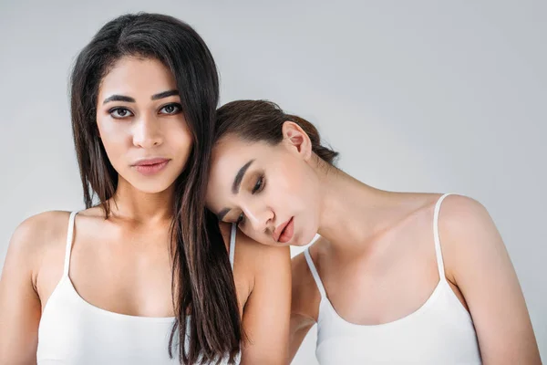 young woman holding head on shoulder of multiracial attractive female friend isolated on gray background