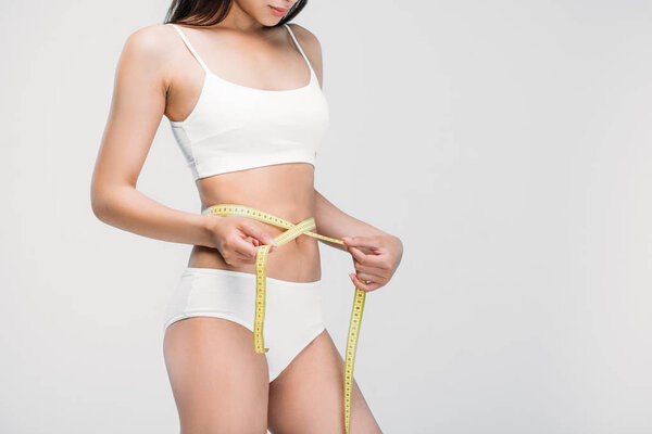 cropped view of slim girl in underwear measuring her waistline, isolated on grey
