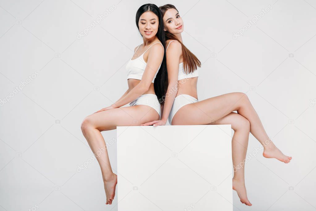 beautiful multicultural girls in underwear posing on white cube, isolated on grey