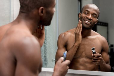 smiling african american man shaving with electric trimmer in bathroom clipart