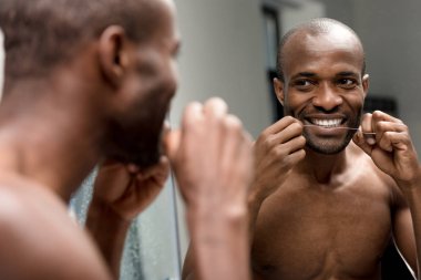 Diş ipi tutan ve banyo aynaya bakarak Afrikalı-Amerikalı genç gülümseyen