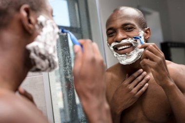 handsome smiling african american man shaving and looking at mirror clipart