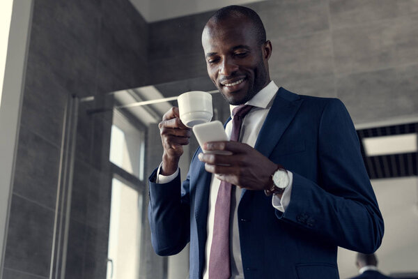 bottom view of smiling young businessman with cup of coffee using smartphone in bathroom