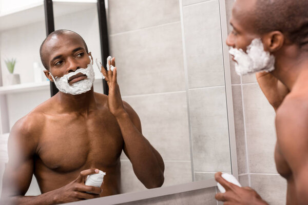 handsome young man applying shaving gel while looking at mirror in bathroom