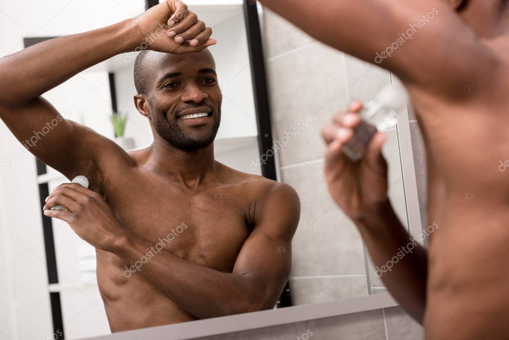 smiling bare-chested african american man applying antiperspirant in bathroom