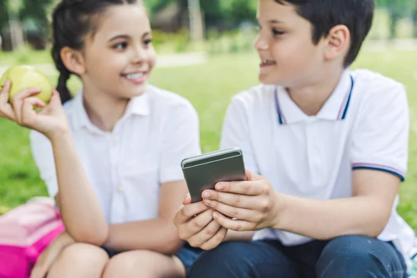 スマート フォンを併用し ながら草の上に座ってチャット小学生 — ストック写真