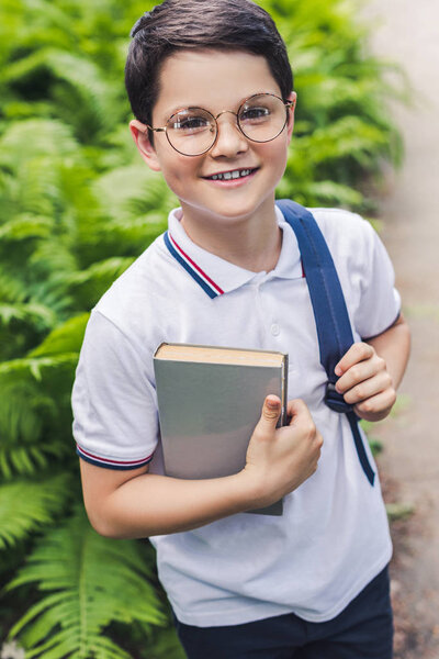 happy schoolboy with backpack and book looking at camera in garden