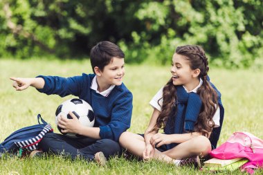 mutlu okul sırt çantaları ve futbol topu ile parkta çim üzerinde oturan ve sohbet