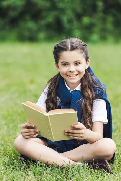 微笑的女孩与书坐在草在公园和看照相机 — 图库照片