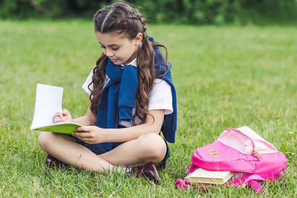 smiling schoolgirl reading book while sitting on grass in park