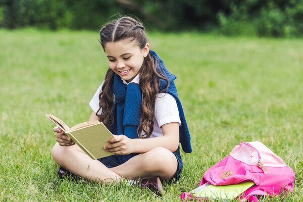 happy schoolgirl reading book while sitting on grass in park
