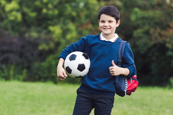 happy kid with soccer ball and backpack showing thumb up on grass field