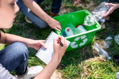cropped view of people cleaning park with green recycling box and writing in textbook clipart