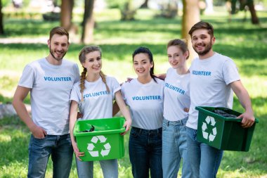 young volunteers with green recycling boxes for trash standing in park  clipart