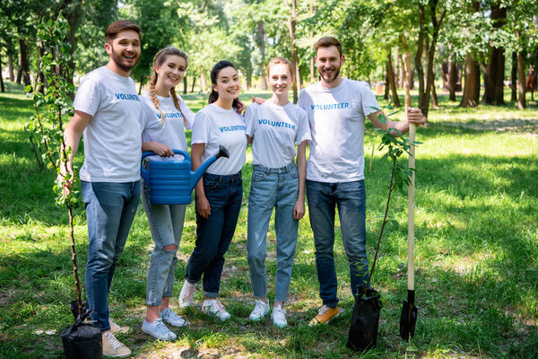 smiling volunteers with new trees, watering can and shovel standing in park  