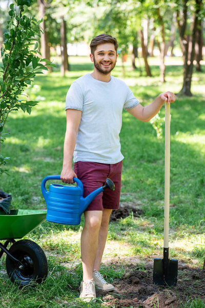 человек с лейкой и лопатой, стоящий в парке с новыми деревьями в тачке
