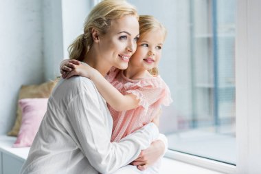 beautiful happy mother and daughter hugging and looking at window clipart