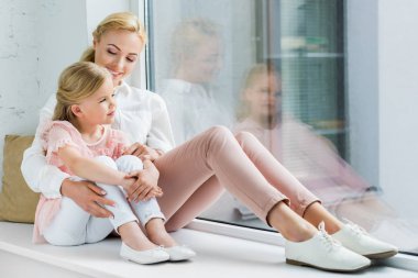 beautiful happy mother and daughter sitting together on windowsill clipart