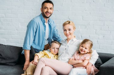 iki çocukla birlikte kanepede otururken kameraya gülümseyen mutlu aile