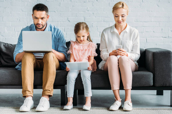 family with one child sitting on couch and using digital devices 