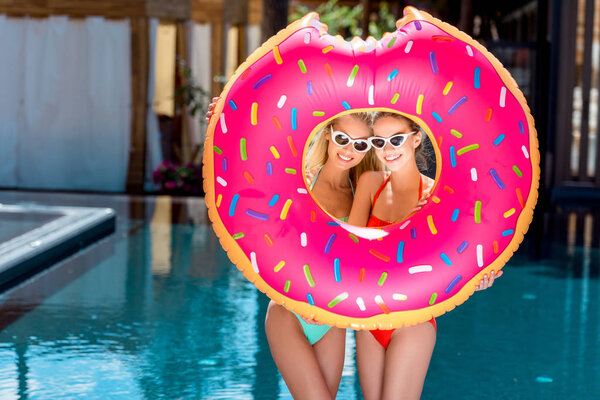 beautiful young women looking at camera through inflatable ring in shape of bitten donut at poolside