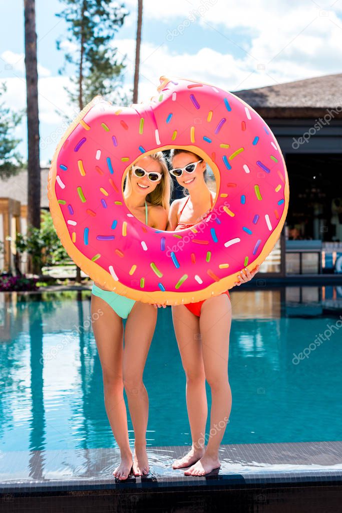 smiling young women looking at camera through inflatable ring in shape of bitten donut at poolside