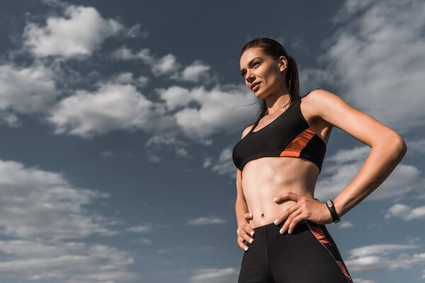 attractive sportswoman posing with hands on hips against blue cloudy sky