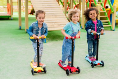 üç çok kültürlü çok güzel küçük çocuklar üzerinde sürme scooter oyun alanı tekme. 