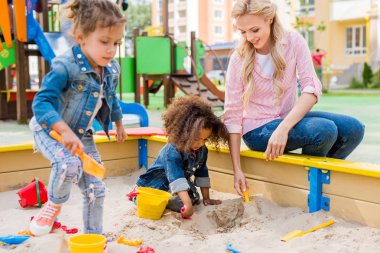 selective focus of mother playing with daughter and her little friend in sandbox at playground  clipart