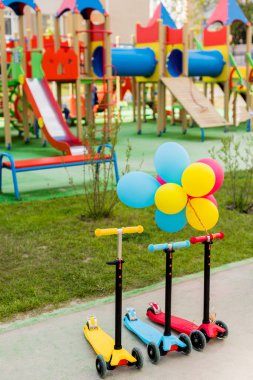 three childish kick scooters placed in row with colorful air balloons at playground  clipart