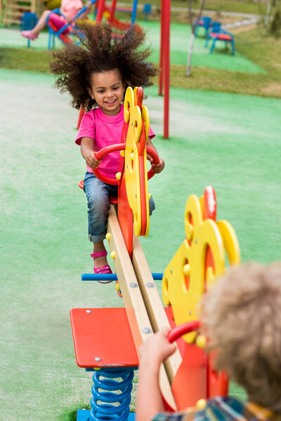 happy curly african american child riding on rocking horse with boy at playground 
