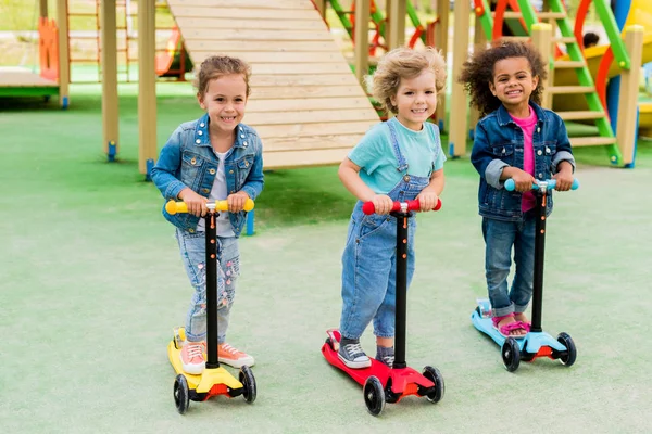 Drei Multikulturell Liebenswerte Kleine Kinder Auf Tretrollern Auf Spielplatz — Stockfoto