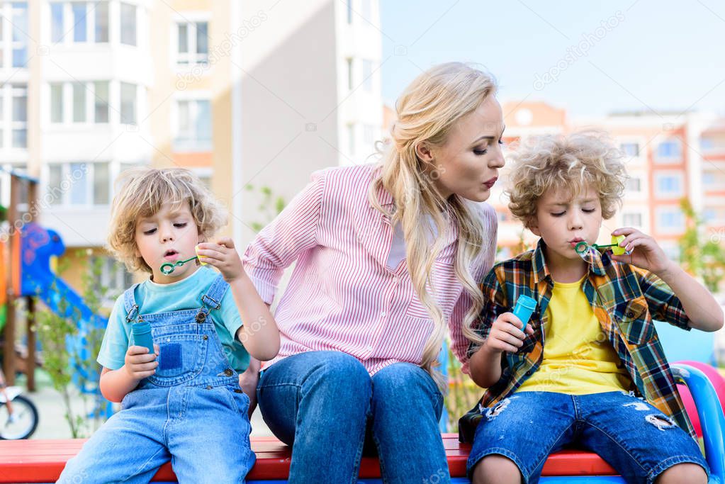 selective focus of mother sitting between two adorable little sons while they using bubble blowers on bench