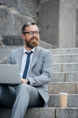 dizüstü bilgisayar kullanarak ve tek kullanımlık fincan kahve ile merdivenlerde oturan yakışıklı işadamı