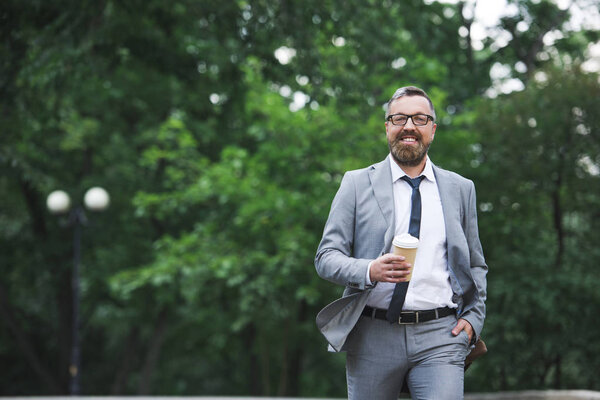 bearded smiling businessman walking on street and holding disposable coffee cup